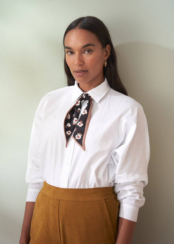 Woman wearing a white shirt with a floral scarf and brown cord trousers against a plain background