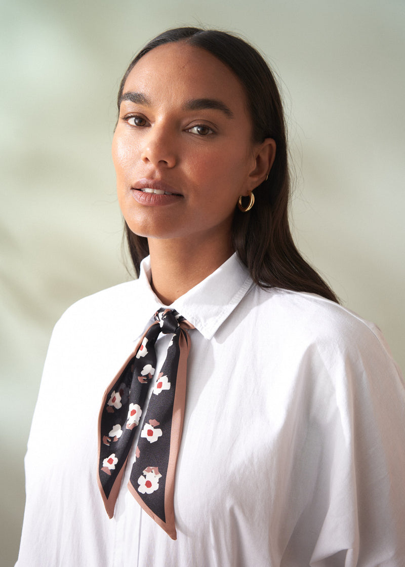Woman wearing a white shirt with a floral scarf around her neck against a plain background