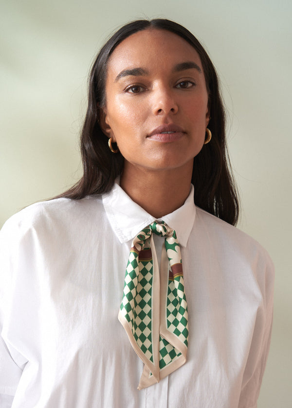Woman wearing a white shirt with a green and beige checkered scarf around her neck against a plain background