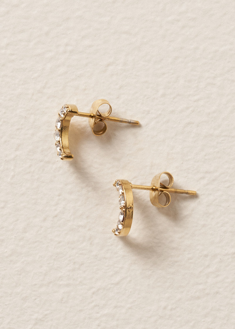 Gold moon stud earrings with embedded crystals on a beige textured background