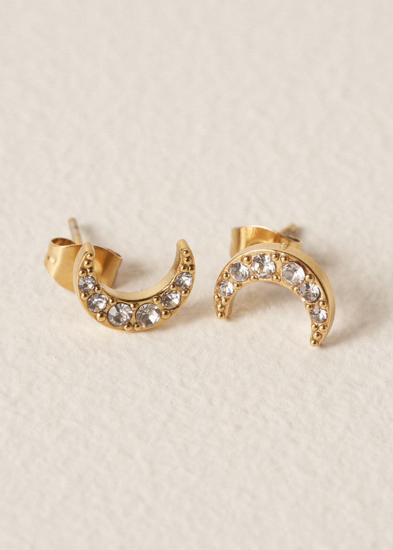 Gold moon stud earrings with embedded crystals on a beige textured background