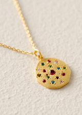 A gold pendant necklace featuring a round, textured gold medallion adorned with small, colorful gemstones in red, green, blue, and clear, suspended from a delicate gold chain on a textured off-white background.