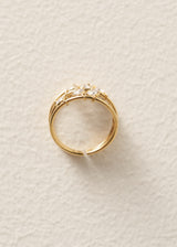 A gold open-band ring with a delicate floral crown design and clear gemstones, photographed on a textured off-white background.