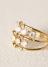 A gold bangle-style ring with multiple clear round gemstones in bezel settings, photographed close-up on a soft light beige background.
