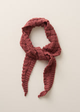 A textured, chunky-knit scarf in a dusty rose or muted terracotta color, styled in a loose knot on a plain, warm off-white background.
