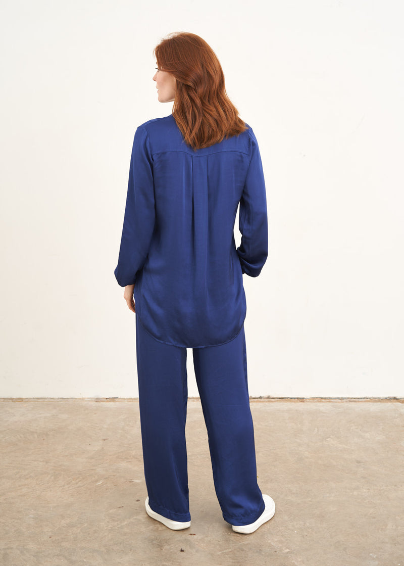 A model wearing a navy blue satin co-ord consisting of a long sleeved blouse with a v neck and wide leg long trousers