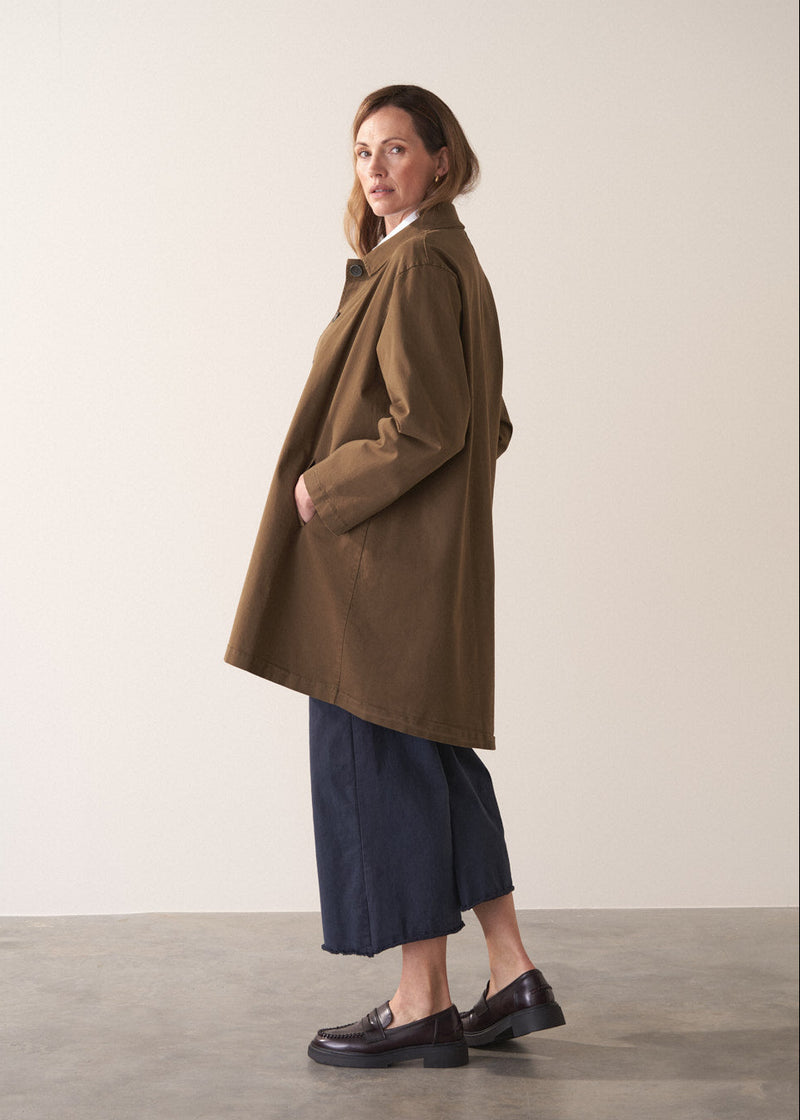 Woman wearing a khaki brown short trench coat over a white shirt on a plain background