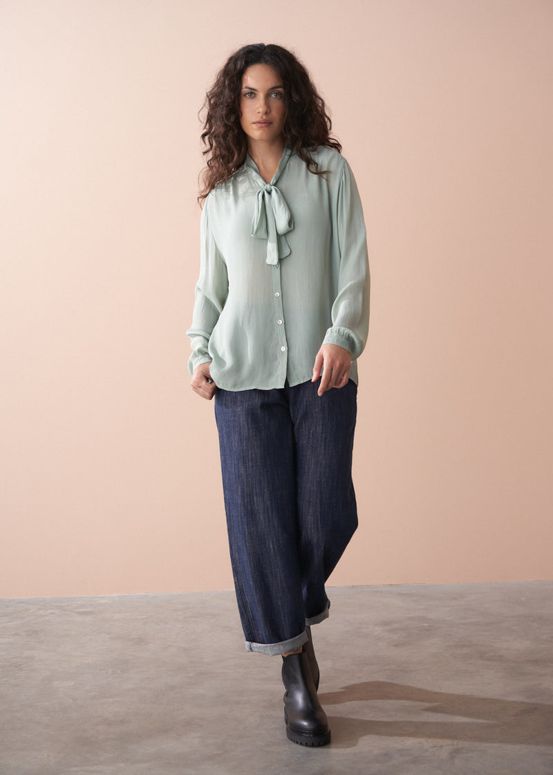 Woman wearing a light green blouse with a tie and dark blue pants against a beige background