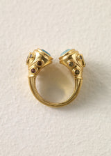 A gold open ring with two decorative ends featuring light aqua semi precious oval stones and small dark brown gem accents, photographed on a textured off-white background