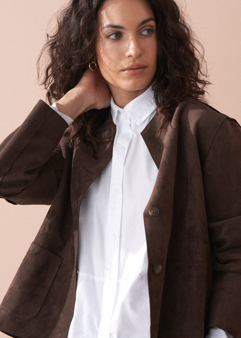 Woman wearing a brown faux suede jacket over a white shirt with a beige background