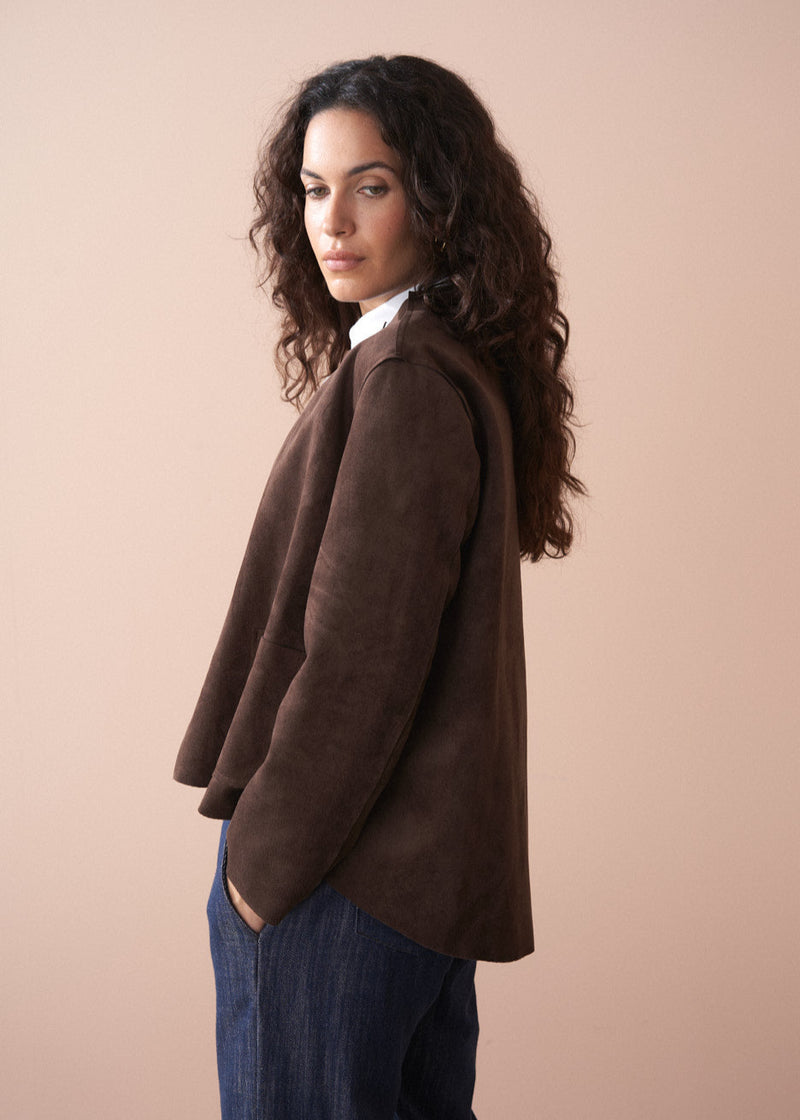 Woman wearing a brown faux suede jacket over a white shirt with a beige background