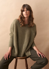 Woman wearing a khaki green wool blend sweater and pants sitting on a wooden stool against a beige background