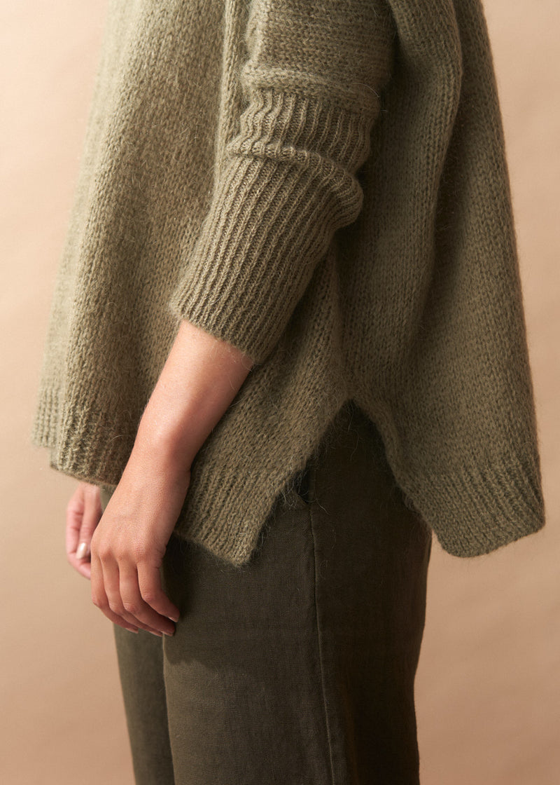 Person wearing a khaki green wool blend knitted sweater and pants against a beige background