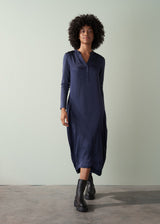 Woman in a long, dark navy v-neck satin dress and black leather boots, standing cross legged against a plain green backdrop