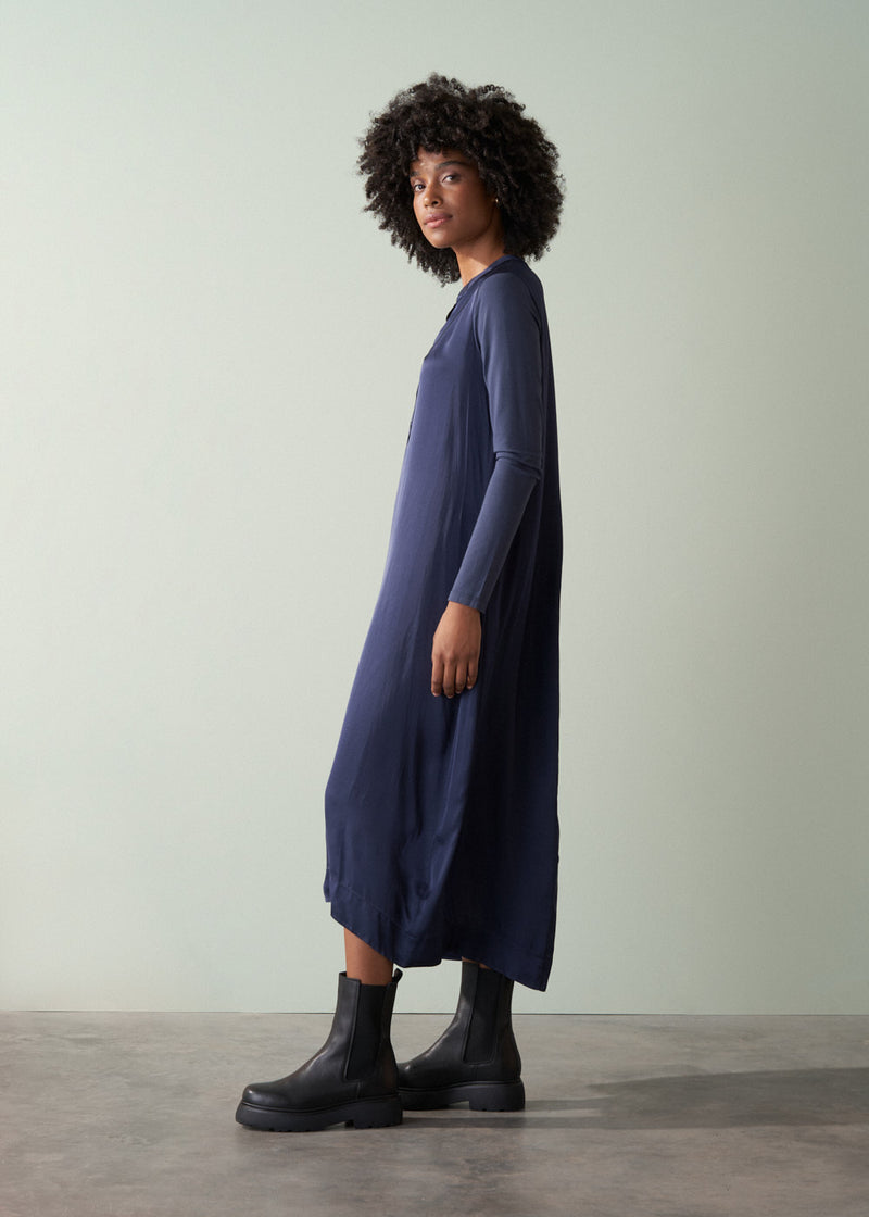 Woman in a long, dark navy satin dress and black leather boots against a plain background