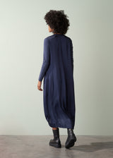 Woman wearing a long, dark navy satin dress and black leather boots facing a plain green wall.