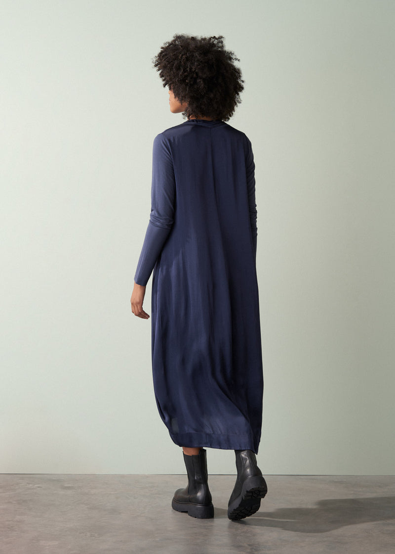 Woman wearing a long, dark navy satin dress and black leather boots facing a plain green wall.