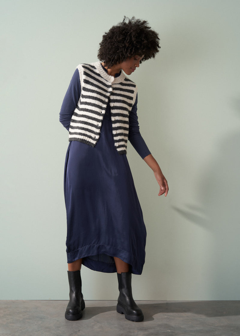 A woman in a long, dark navy satin dress, striped knitted waistcoat, and black leather boots pointing at the floor.