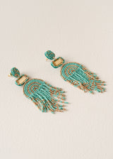 A pair of beaded tassel earrings featuring turquoise and gold seed beads in a semi-circular arch pattern with dangling fringe, displayed on a textured off-white background.