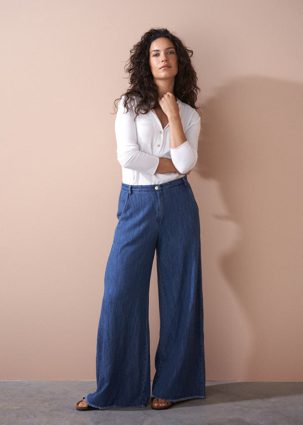 Woman wearing a white blouse and blue wide-leg pants against a beige wall.