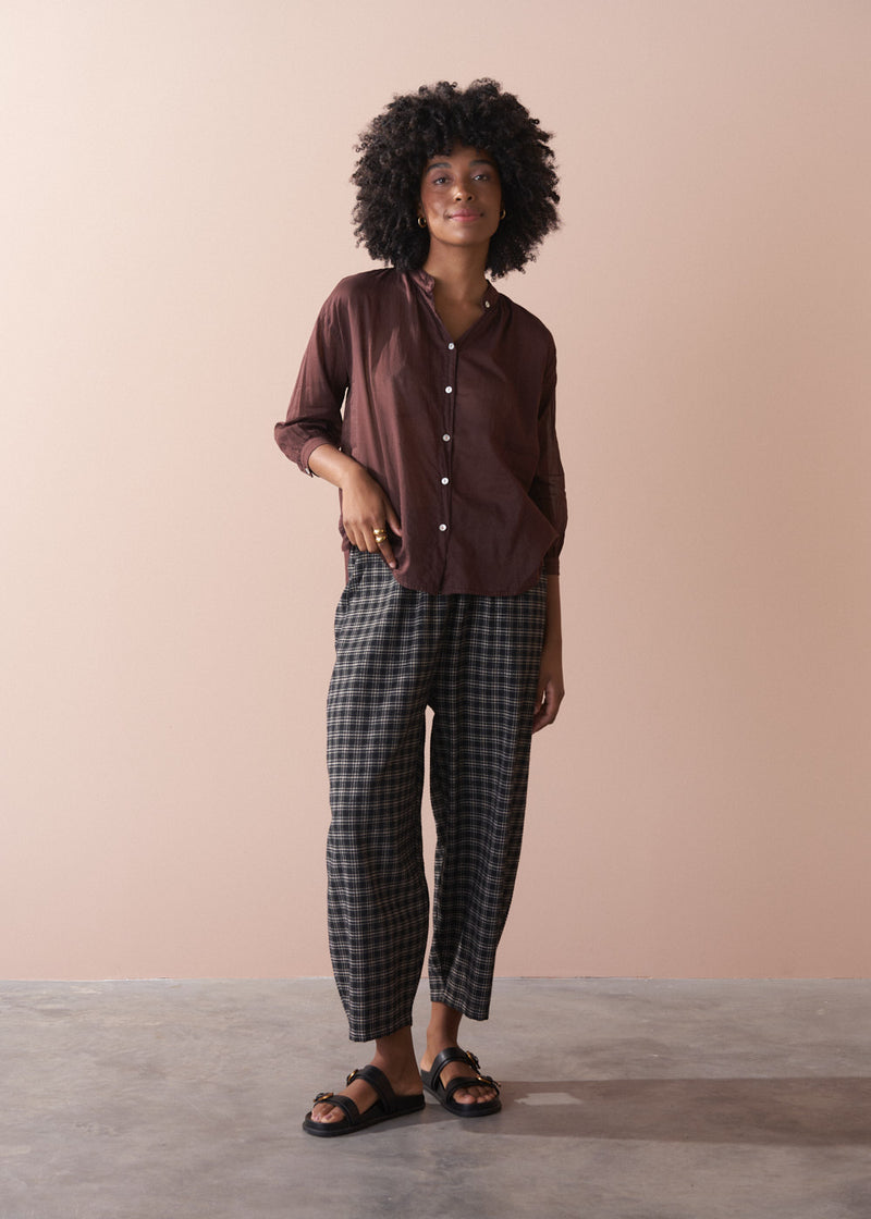 Woman wearing a brown semi sheer shirt and checkered pants against a beige background