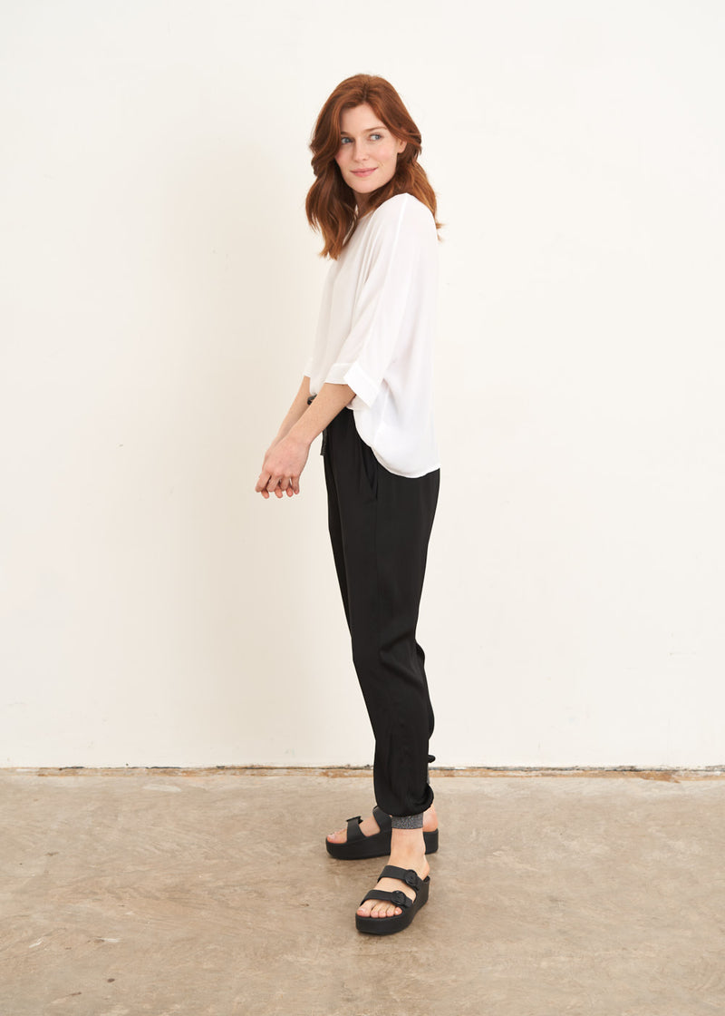 A model wearing a pair of black satin trousers with silver glitter details on the cuffs and tie waist with a white slouchy top and black slides