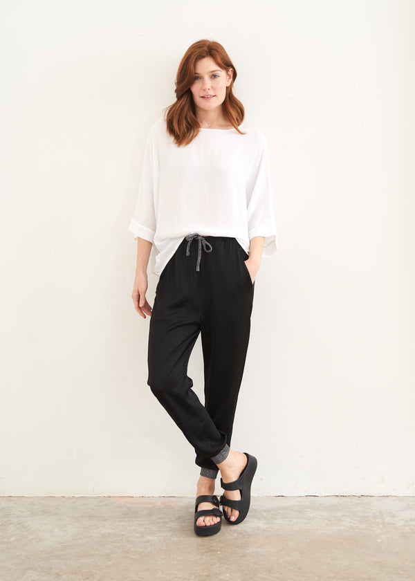 A model wearing a pair of black satin trousers with silver glitter details on the cuffs and tie waist with a white slouchy top and black slides