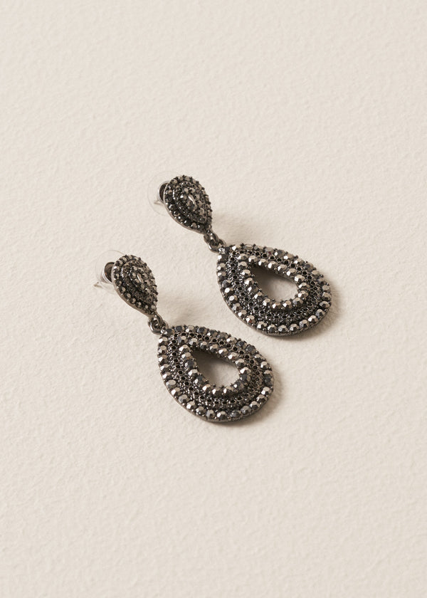A pair of Ainsley Earrings featuring a striking teardrop silhouette embellished with jet-black crystals, on a beige background