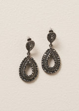 A pair of Ainsley Earrings featuring a striking teardrop silhouette embellished with jet-black crystals, on a beige background