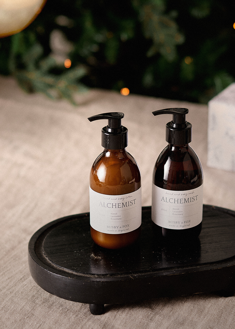 Hand and body wash in an amber bottle with black pump and "Alchemist" branding, on a wooden stand. 