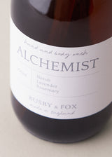 Close up of hand and body wash in an amber bottle with black pump and "Alchemist" branding, on a beige background
