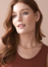 Woman wearing a gold necklace with heart-shaped charms, styled with a brown jersey vest.