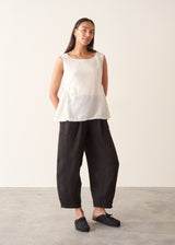 A woman wearing a sleeveless white top with a relaxed, slightly sheer texture and a peplum-like hem, paired with dark wide-leg trousers.