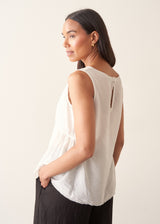 A woman wearing a sleeveless white top with a relaxed, slightly sheer texture and a peplum-like hem, paired with dark wide-leg trousers.