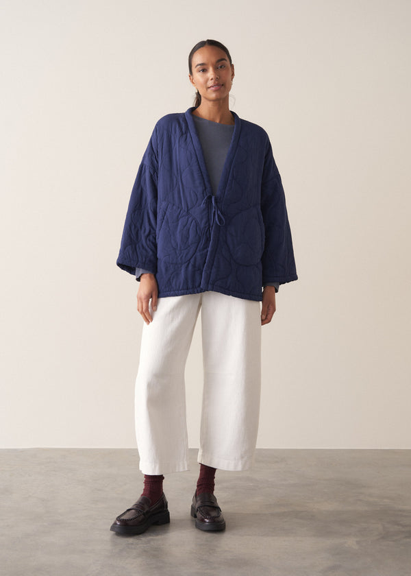 Person wearing a navy blue jacket and white barrel leg trousers against a plain background