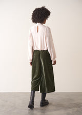 Woman wearing green velvet cropped wide-leg trousers and a pink shirt against a plain background