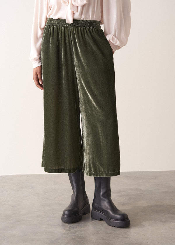 Woman wearing green velvet cropped wide-leg trousers and a pink shirt against a plain background