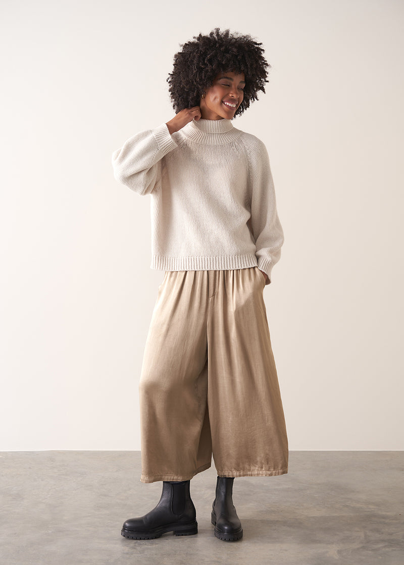 Woman wearing a white rollneck sweater and beige wide leg trousers against a white background