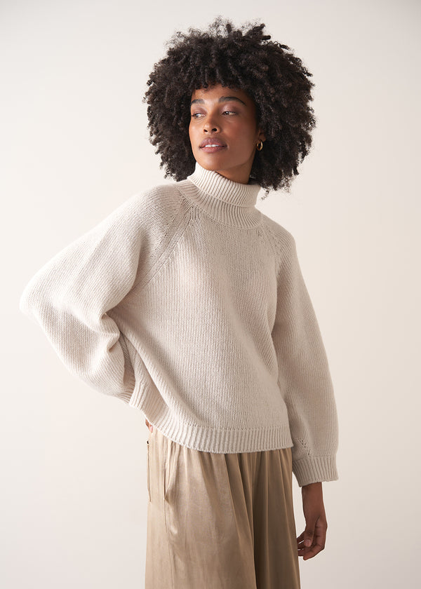 Woman wearing a white rollneck sweater and beige wide leg trousers against a white background