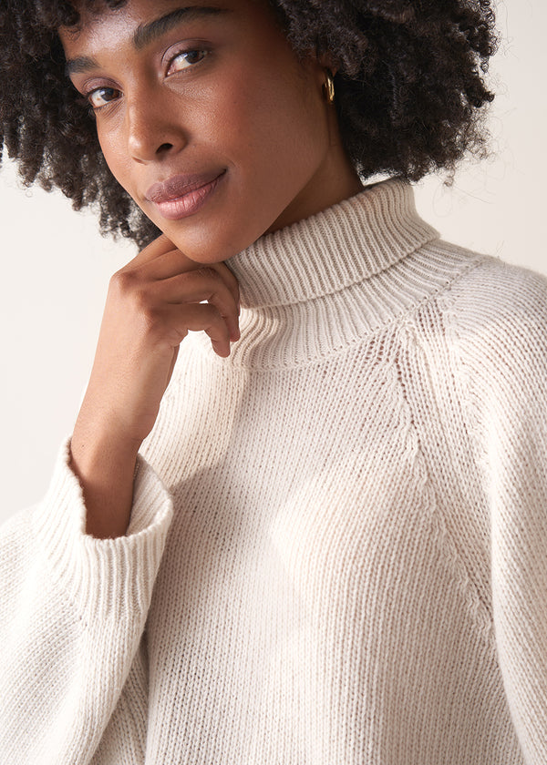 Woman wearing a white rollneck sweater and beige wide leg trousers against a white background