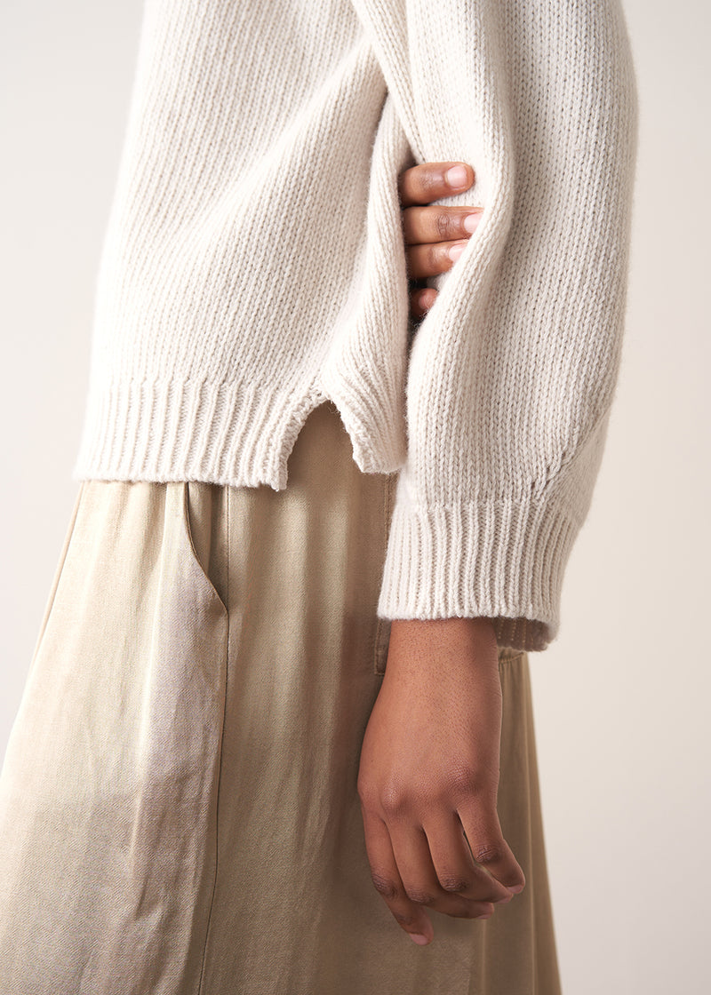 Woman wearing a white rollneck sweater and beige wide leg trousers against a white background