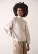 Woman wearing a white rollneck sweater and beige wide leg trousers against a white background