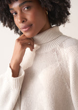 Woman wearing a white rollneck sweater and beige wide leg trousers against a white background