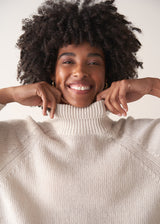 Close up of woman wearing a white rollneck sweater and beige wide leg trousers against a white background