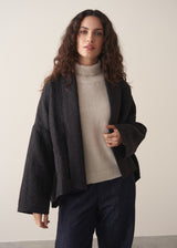 Woman wearing a black quilted kimono jacket over a beige sweater and dark jeans against a plain background