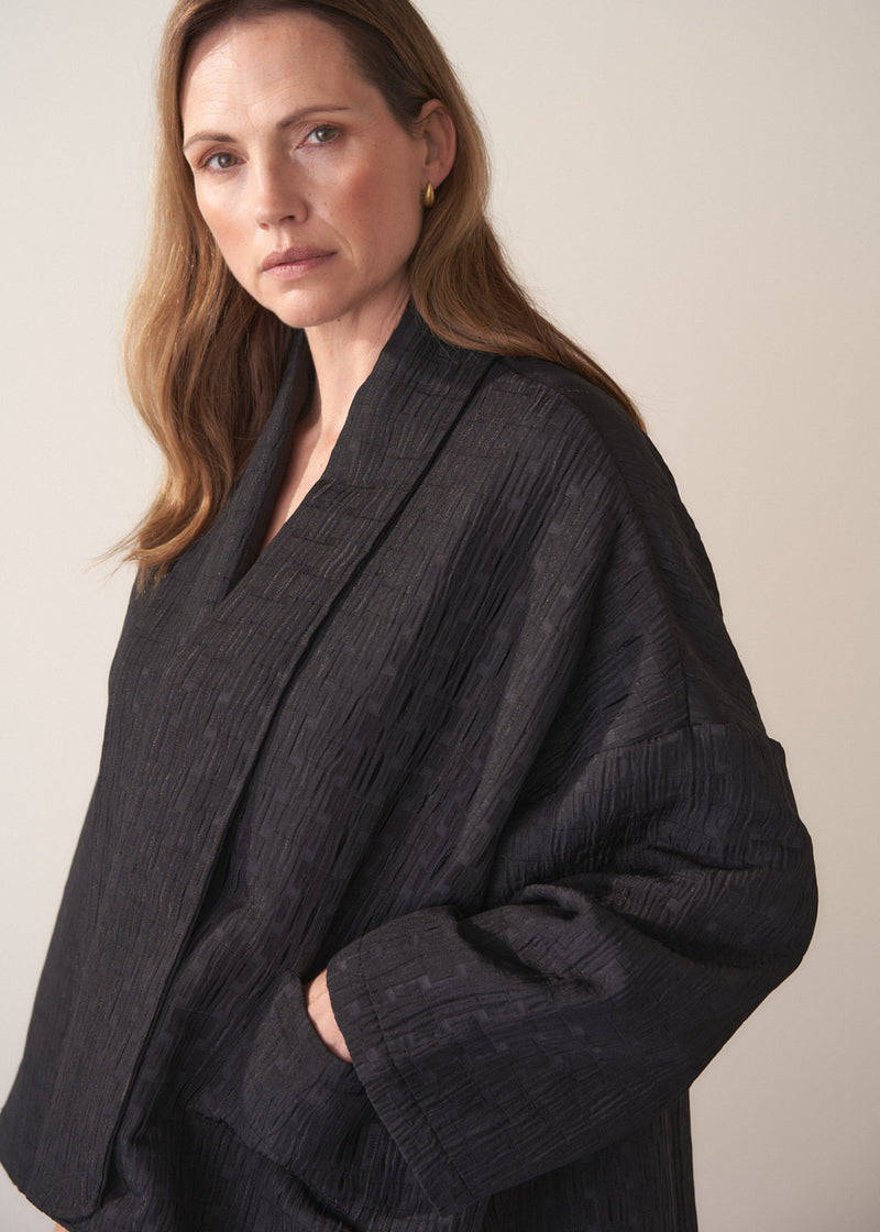 Woman wearing a oversized quilted jacket over a white top and gold trousers against a light background