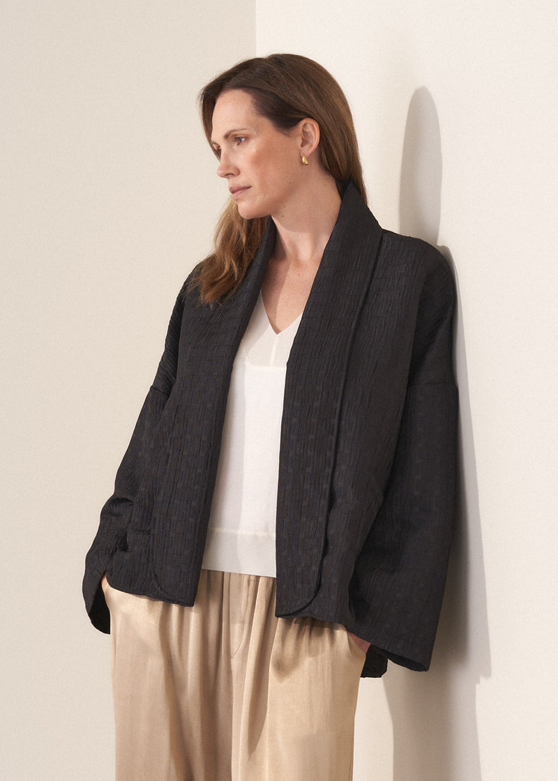 Woman wearing a oversized quilted jacket over a white top and gold trousers against a light background