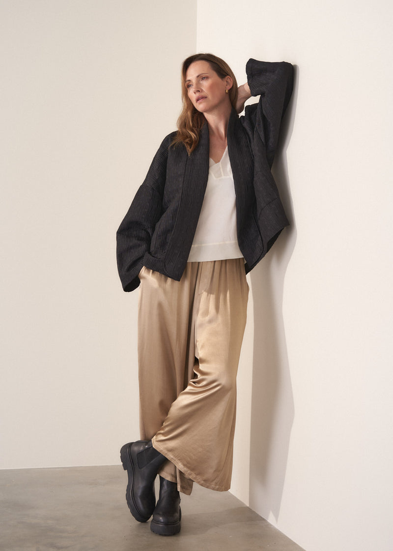 Woman wearing a oversized quilted jacket over a white top and gold trousers against a light background