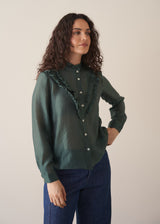 Woman wearing dark green ruffle front shirt with blue jeans against a beige background