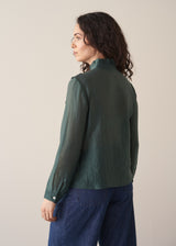 Woman wearing dark green ruffle front shirt with blue jeans against a beige background